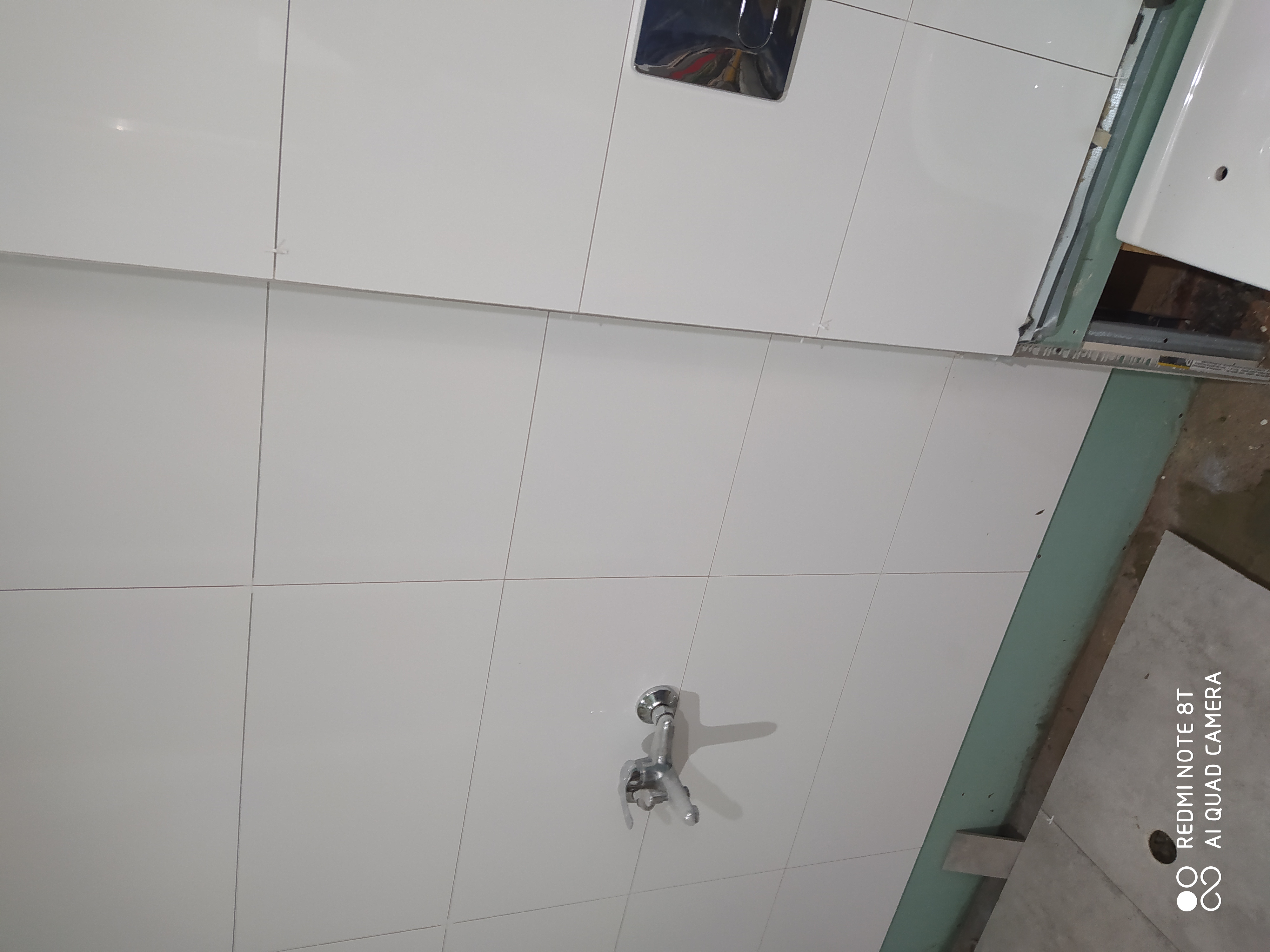 Białe, błyszczące płytki ceramiczne na ścianie łazienki, widoczny fragment wnęki z instalacją wodną i baterią.