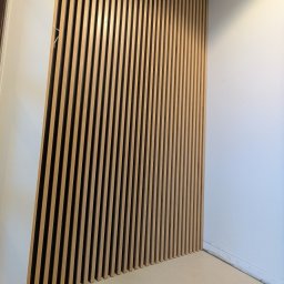 DREAMWOOD SYSTEM Przemysław Pachuta - Drewniana ściana z pionowych listew w jasnym odcieniu, tworząca dekoracyjny panel na tle białej ściany i jasnej podłogi.