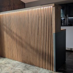 DREAMWOOD SYSTEM Przemysław Pachuta - Drewniana ścianka działowa z pionowych listew, podświetlona od góry, tworząca element dekoracyjny i funkcjonalny w pomieszczeniu z marmurową podłogą i elementami biurowymi w tle.