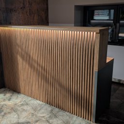 DREAMWOOD SYSTEM Przemysław Pachuta - Drewniana lamelowa recepcja z podświetleniem LED, widok z boku na tle mozaikowej podłogi i ściany z efektem rdzy, obok fotel z poduszką i roślinką doniczkową.