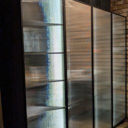 DREAMWOOD SYSTEM Przemysław Pachuta - Przesuwne drzwi do szafy wnękowej z ryflowanego, półprzezroczystego szkła w czarnej ramie, ustawione wzdłuż ceglanej ściany.