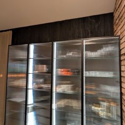 DREAMWOOD SYSTEM Przemysław Pachuta - Cztery moduły przeszklonej szafy z czarnymi ramami, wypełnione naczyniami i dekoracjami, stojące na drewnianej podłodze przy ścianie z ciemnego drewna i cegły.