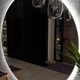 DREAMWOOD SYSTEM Przemysław Pachuta - Owalne lustro z podświetleniem LED w nierównym, teksturowanym obramowaniu, odbijające fragment wnętrza z czarną szafą, drewnianymi stołkami i wiszącymi szklanymi lampami o organicznym kształcie.