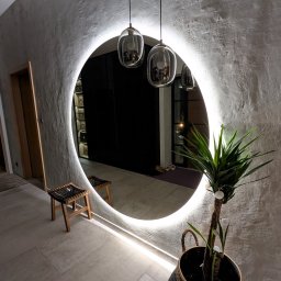DREAMWOOD SYSTEM Przemysław Pachuta - Okrągłe lustro z podświetleniem LED na teksturowanej ścianie, odbijające wnętrze z drewnianym stołkiem i wiszącymi szklanymi lampami, obok doniczka z rośliną.