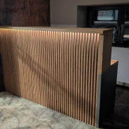 Recepcja z drewnianymi lamelami podświetlonymi od góry, obok skórzany fotel z rośliną doniczkową, w tle okno z biurowymi akcesoriami.