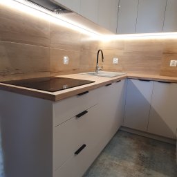 DREAMWOOD SYSTEM Przemysław Pachuta - Nowoczesna kuchnia w kształcie litery L z białymi, matowymi frontami szafek, drewnianym blatem i płytą indukcyjną, oświetleniem LED podszafkowym oraz drewnopodobną okładziną ściany.