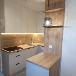 DREAMWOOD SYSTEM Przemysław Pachuta - Nowoczesna kuchnia z białymi, błyszczącymi frontami szafek i drewnianym blatem, oświetlona listwą LED pod szafkami wiszącymi i ozdobną lampą wiszącą z ażurowym kloszem, z widoczną płytą indukcyjną...