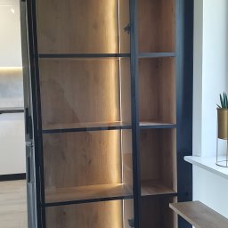 DREAMWOOD SYSTEM Przemysław Pachuta - Półki z drewna z czarnymi metalowymi ramami, oświetlone od tyłu, widoczne zawiasy, na tle kuchni i okna z rośliną w złotej doniczce.