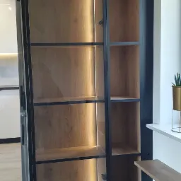 Półki z drewna z czarnymi metalowymi ramami, oświetlone od tyłu, widoczne zawiasy, na tle kuchni i okna z rośliną w złotej doniczce.