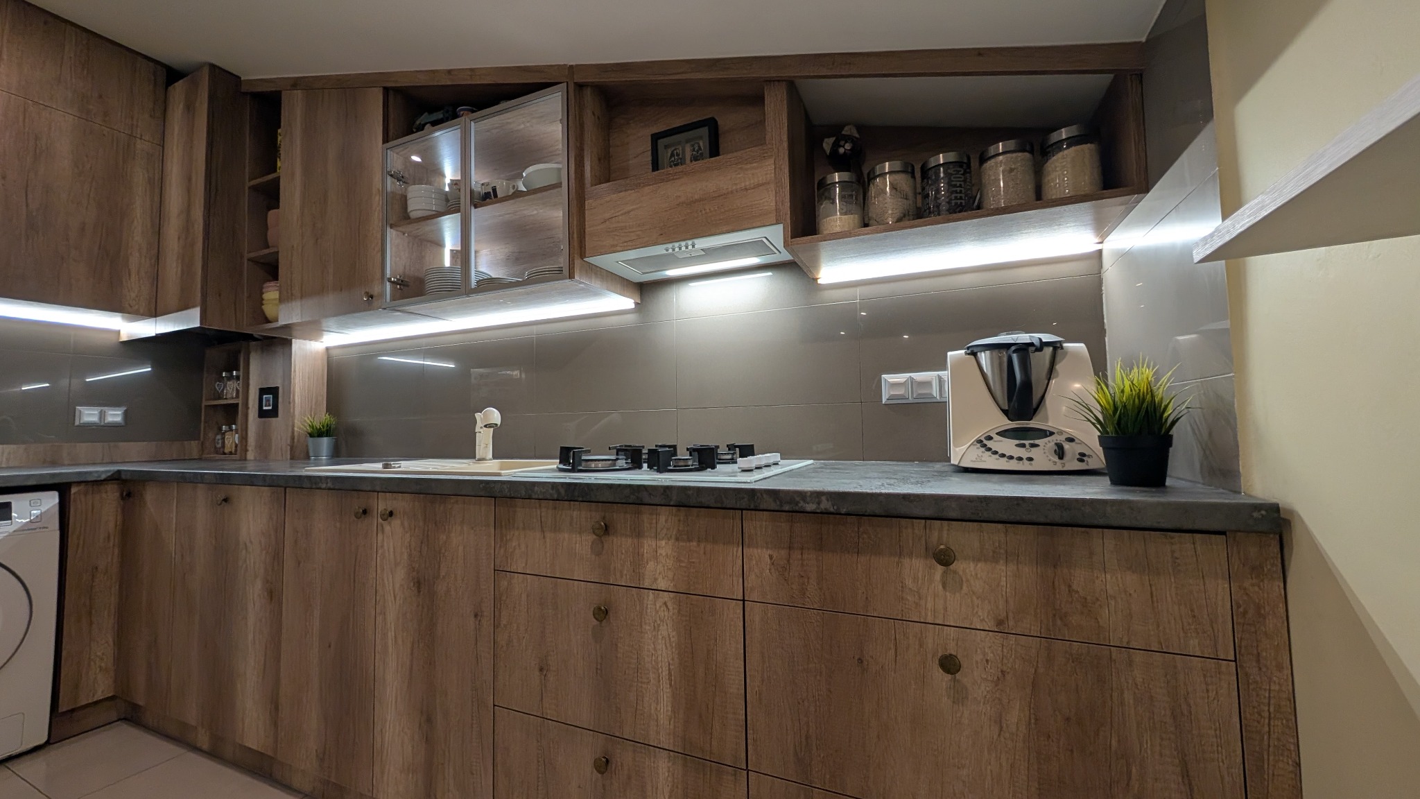 Nowoczesna kuchnia z drewnianymi frontami szafek, szarym blatem i podświetleniem LED pod szafkami wiszącymi, wyposażona w płytę gazową, zlew z baterią i biały robot kuchenny Thermomix na blacie.