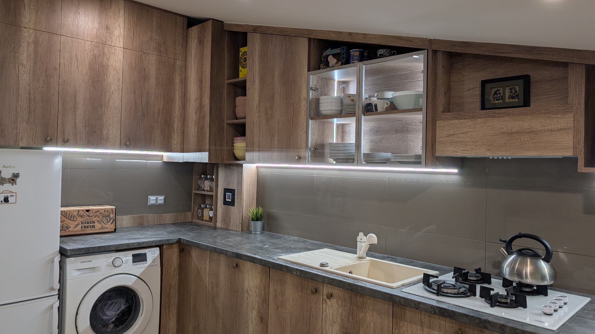 Nowoczesna kuchnia z frontami szafek w odcieniach drewna, oświetleniem LED pod szafkami wiszącymi, szarą glazurą i białymi sprzętami AGD, w tym pralką i lodówką.