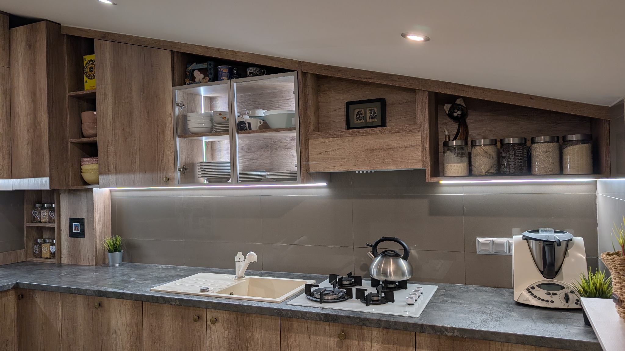 Kuchenna zabudowa z drewnopodobnymi frontami, oświetleniem LED podszafkowym, szklanymi witrynami i skośną szafką, na blacie czajnik, płyta gazowa i robot kuchenny.