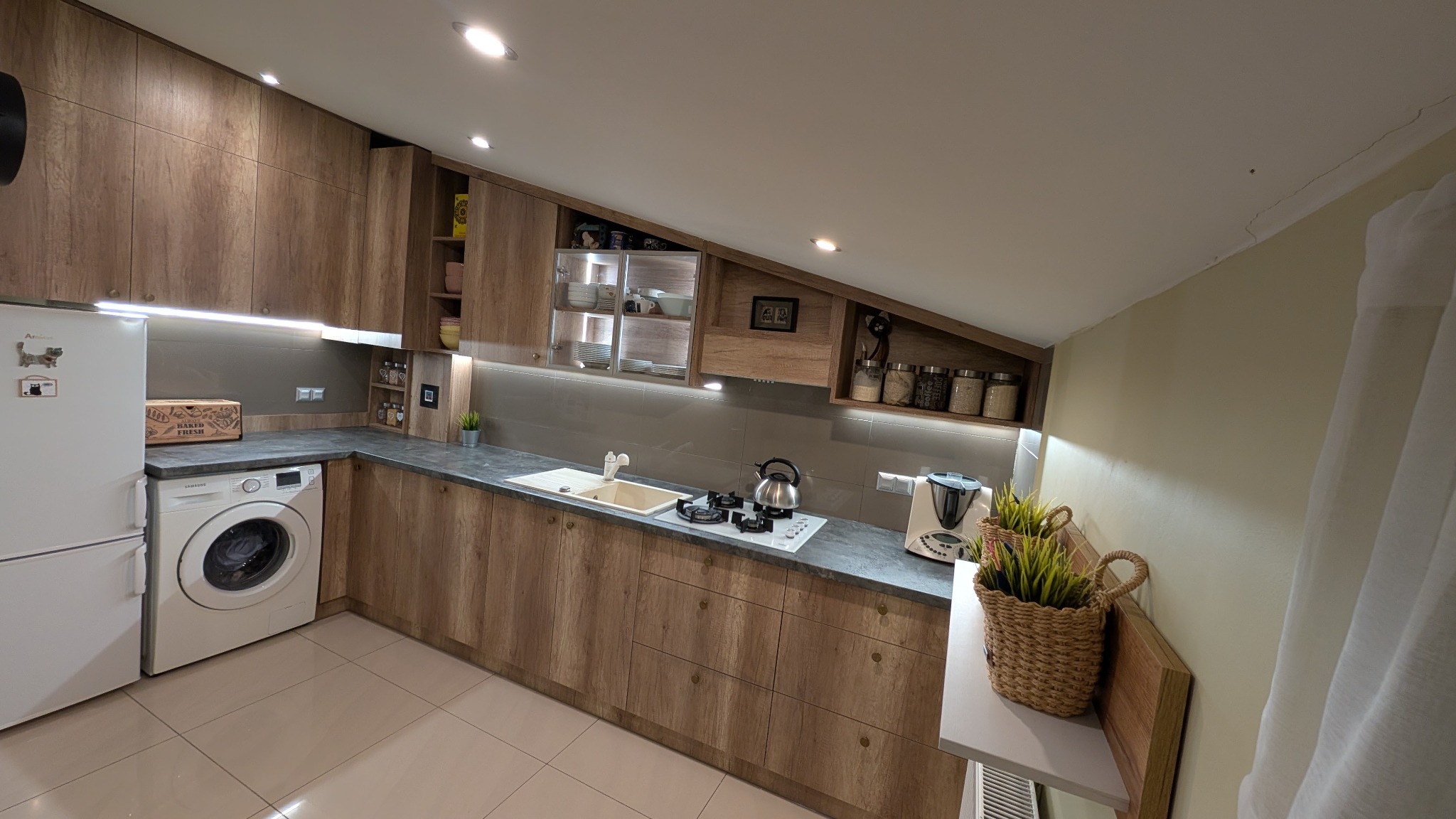 Nowoczesna kuchnia z drewnianymi szafkami, blatem w odcieniach szarości, białymi sprzętami AGD, w tym pralką, oraz dekoracyjnymi roślinami w wiklinowych koszach.