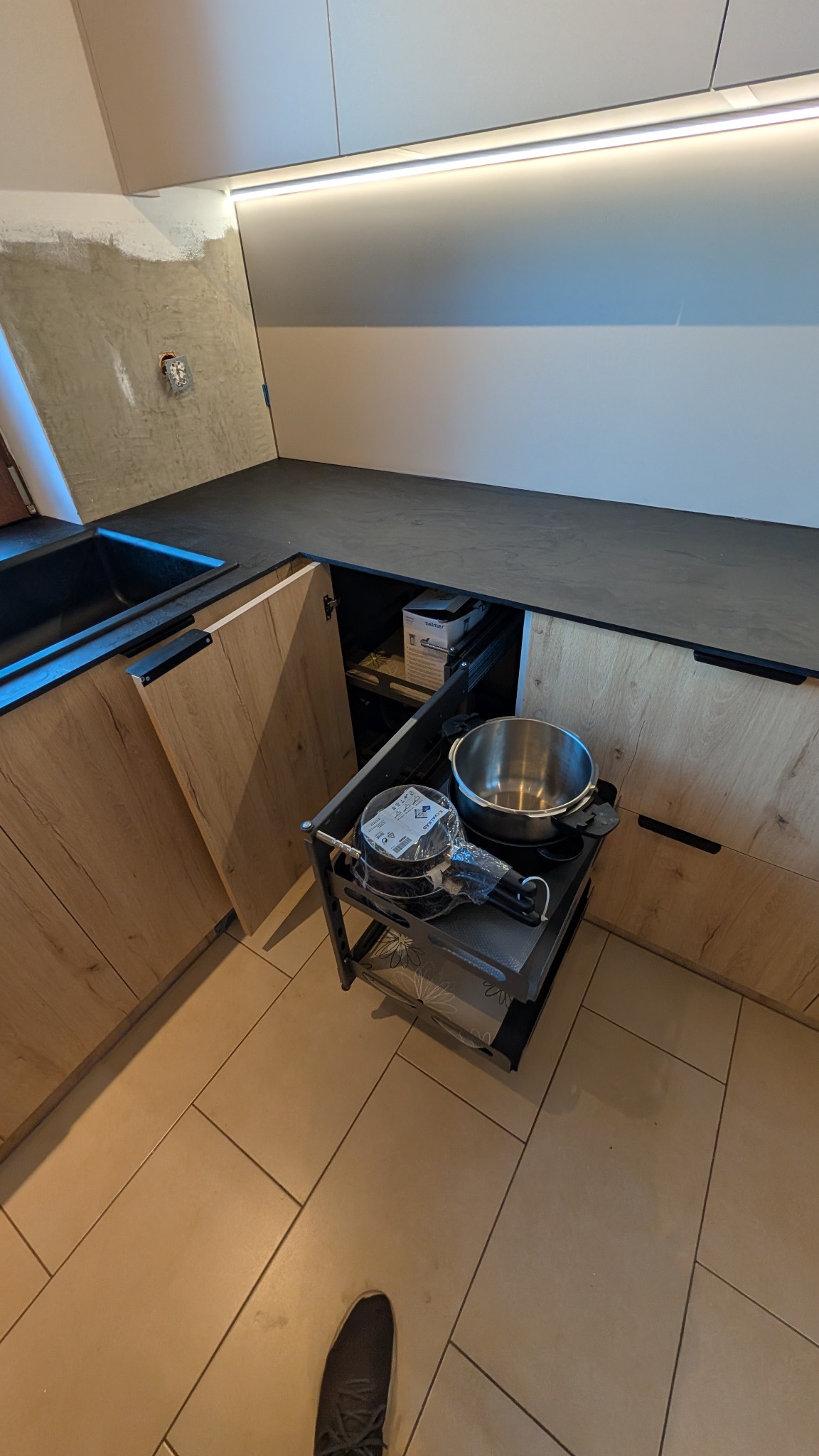 Wnętrze szafki kuchennej z wysuwanym systemem przechowywania garnków, widoczne elementy montażu na ścianie, surowy tynk i fragment buta.