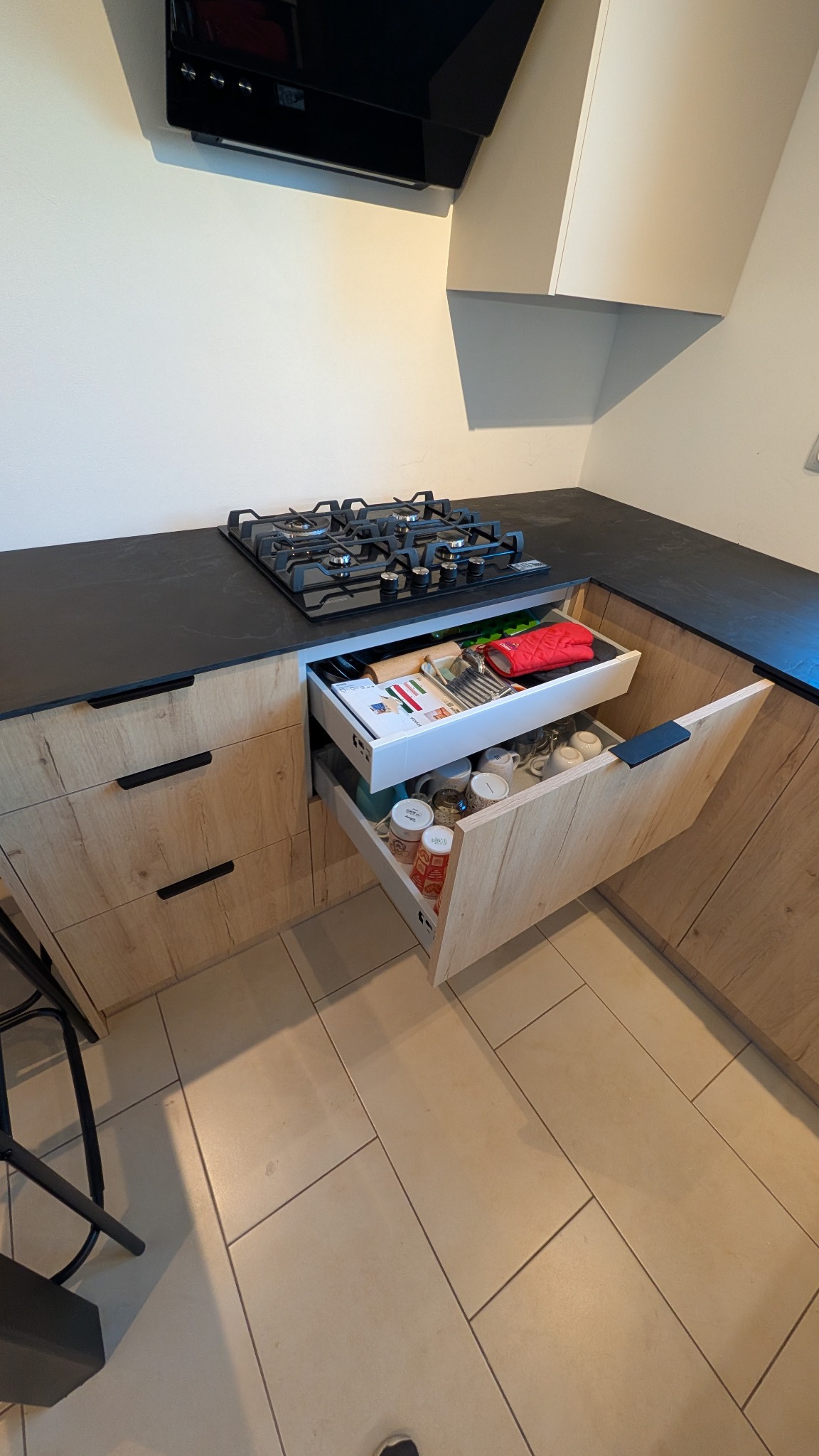 Kuchenna szafka z dwoma wysuwanymi szufladami, górna z sztućcami i akcesoriami kuchennymi, dolna z kubkami, front szafki z drewnopodobnym wzorem, czarny blat i płyta gazowa w tle.