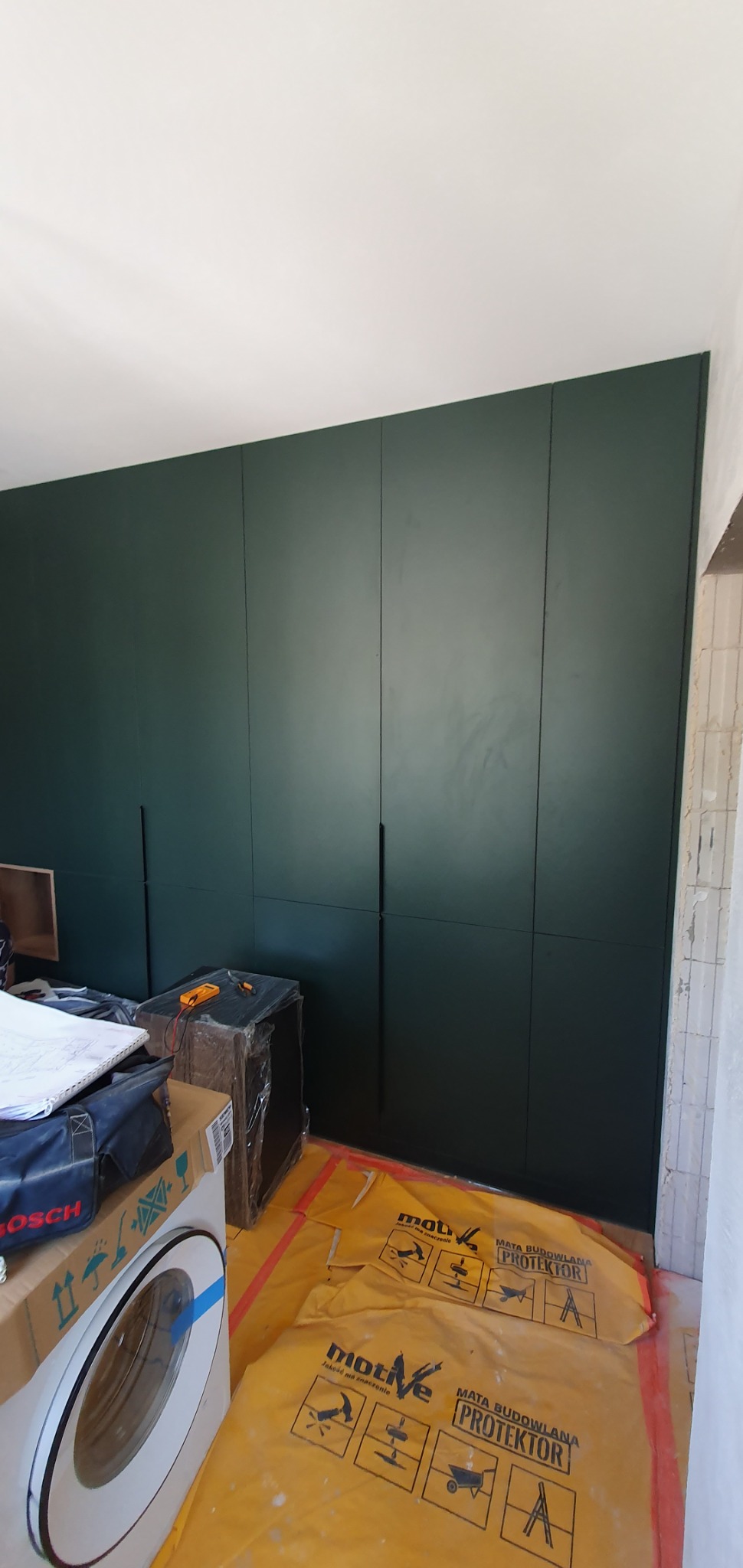 Ciemnozielona szafa wnękowa z długimi, pionowymi uchwytami, widoczna w trakcie montażu; podłoga zabezpieczona folią budowlaną, obok pralka i narzędzia.