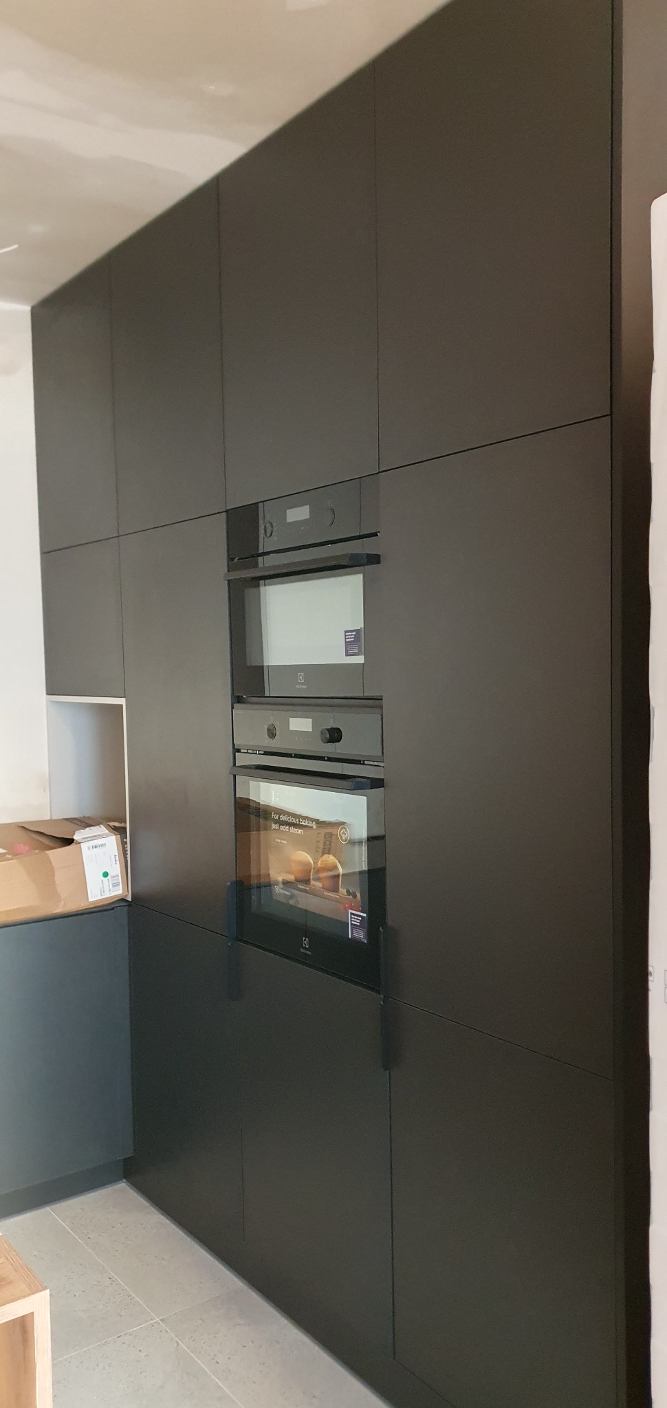 Ciemnoszare szafki kuchenne z wbudowanymi piekarnikami Electrolux, widoczny karton na blacie, surowy stan wykończenia wnętrza.