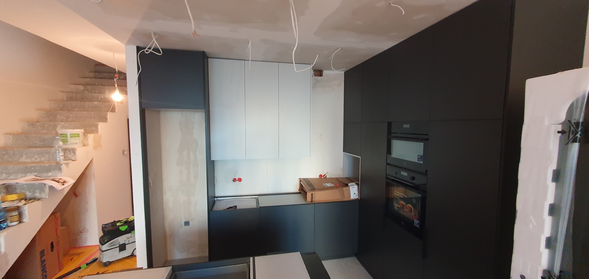 Montaż nowoczesnej kuchni w Lublinie: widoczne białe szafki górne i ciemnoszare dolne, zabudowa piekarnika, instalacja elektryczna w suficie, w tle surowe schody betonowe.