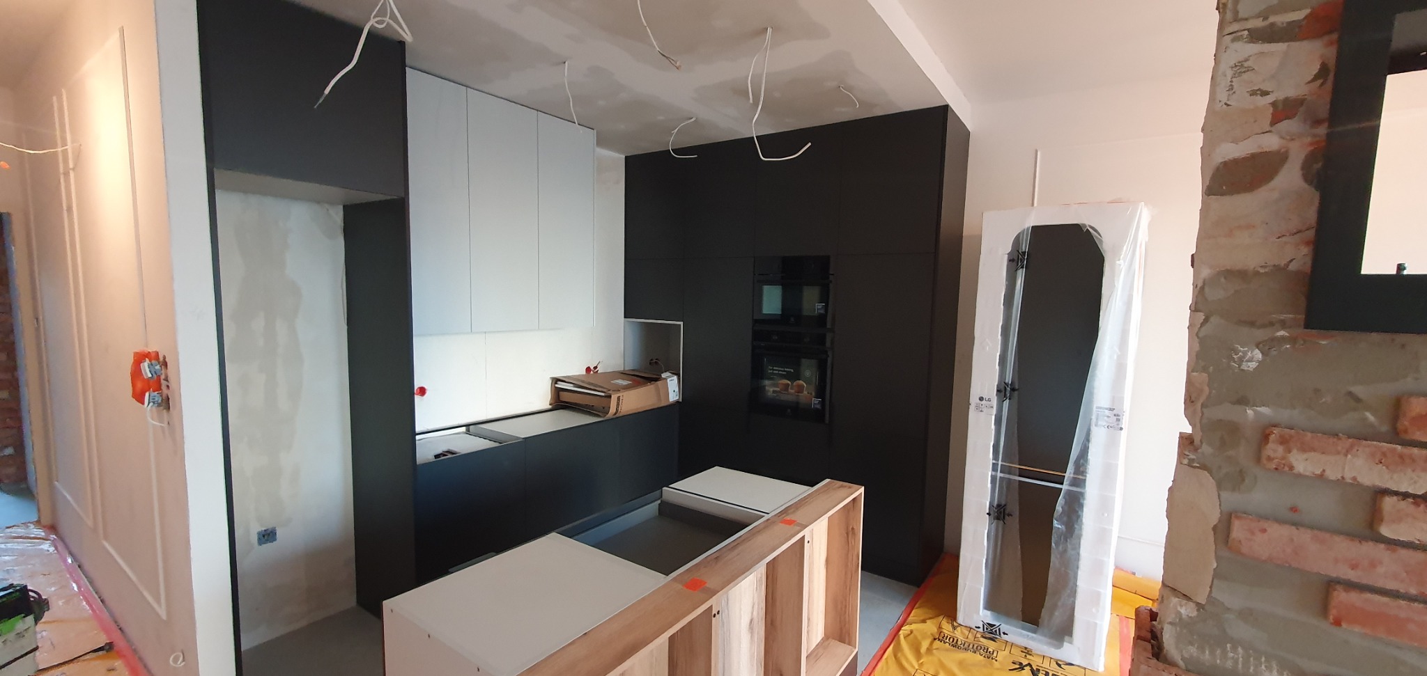 Montaż nowoczesnej kuchni z czarnymi i białymi szafkami, widoczne elementy wyspy kuchennej w surowym drewnie, instalacja elektryczna w trakcie, drzwi w folii ochronnej.