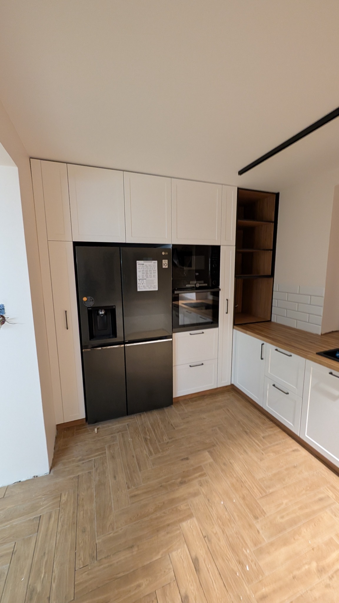 Nowoczesna kuchnia z białymi szafkami, czarną lodówką side-by-side i drewnianym blatem, wykończona podłogą z płytek imitujących parkiet w jodełkę.