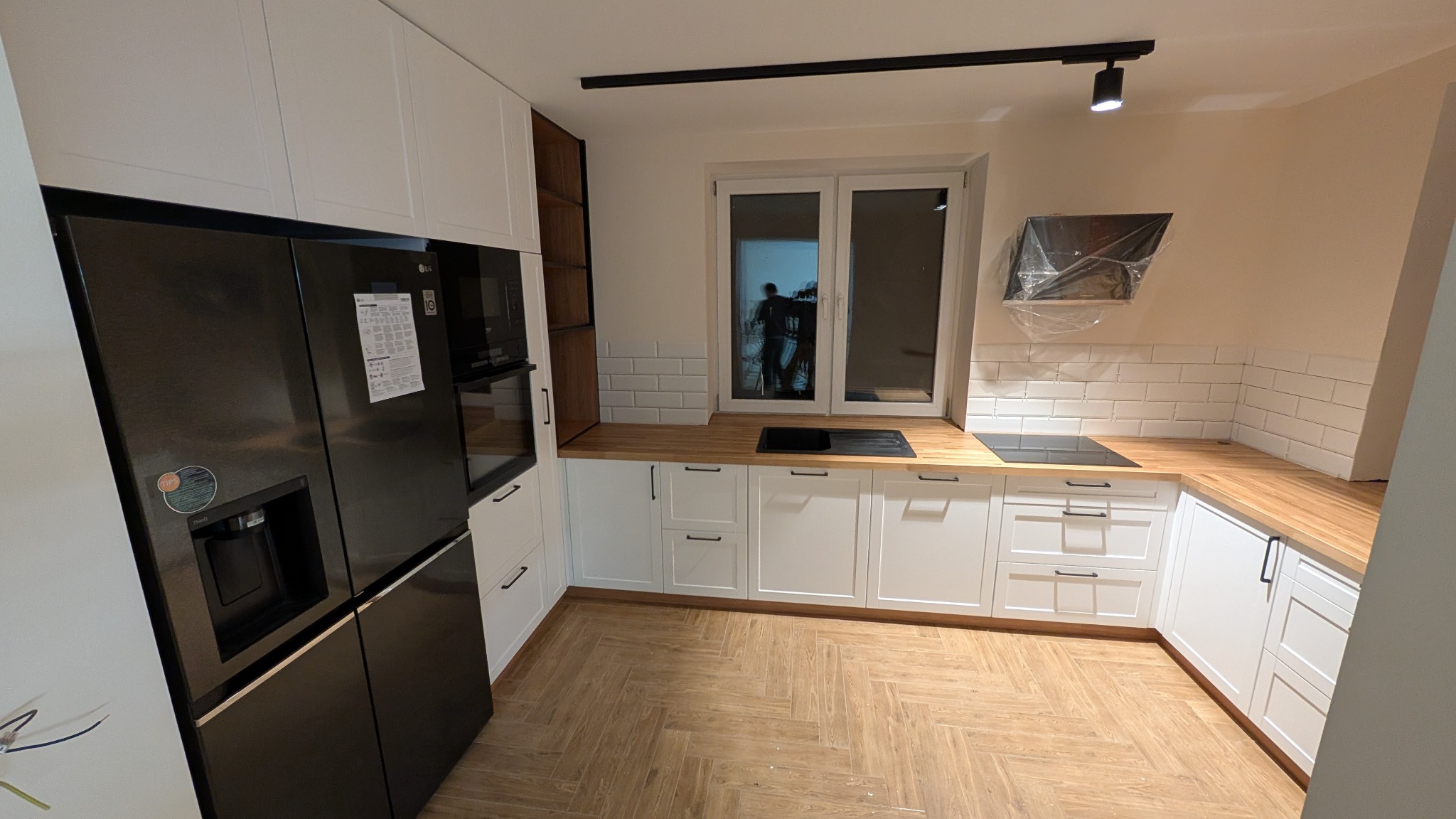 Nowoczesna kuchnia z białymi szafkami, drewnianymi blatami i czarną lodówką side-by-side LG, widoczny okap kuchenny owinięty folią ochronną i osoba w oknie, parkiet ułożony w jodełkę.