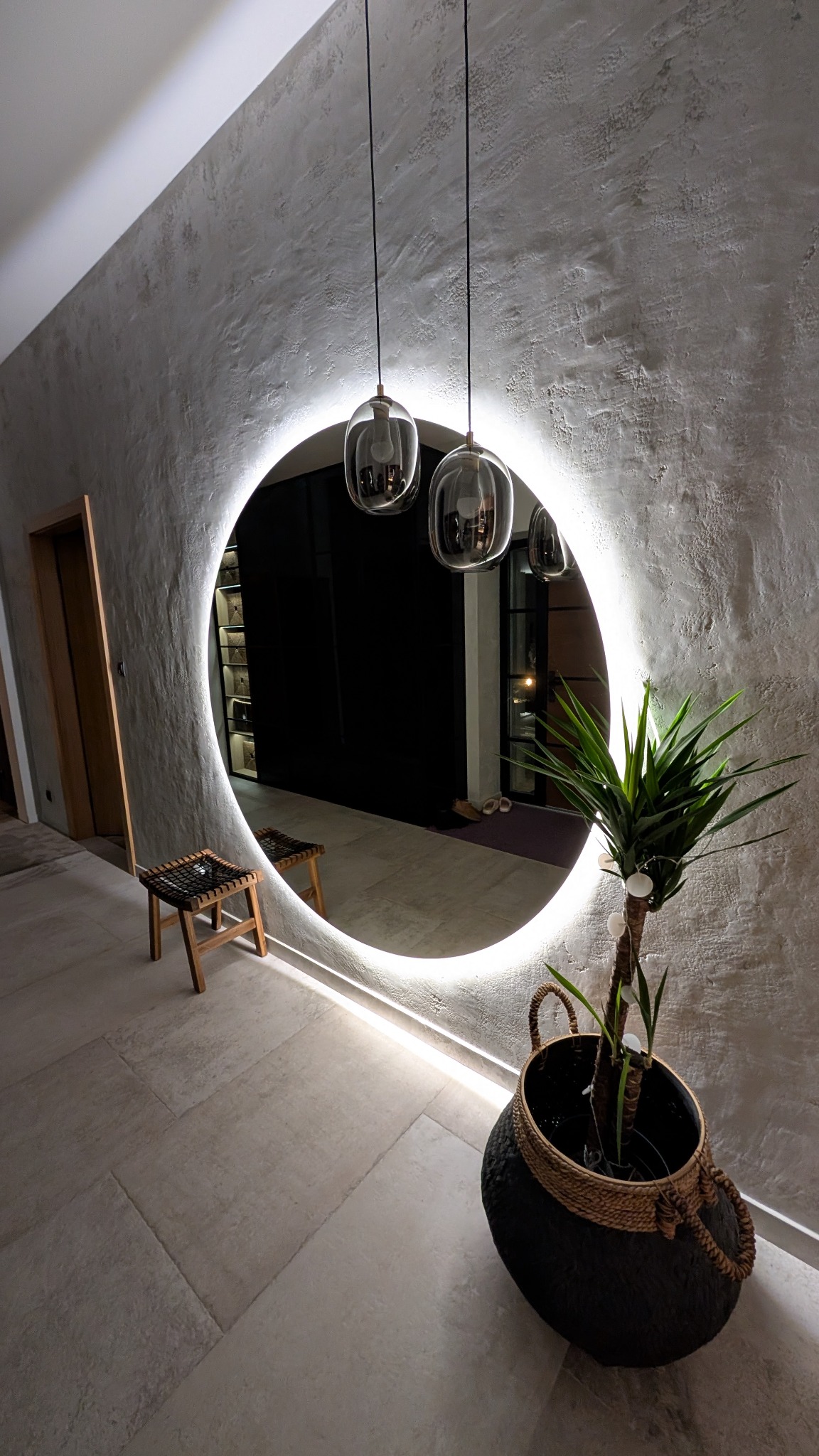 Okrągłe lustro z podświetleniem LED w surowym, teksturowanym wnętrzu, z widocznym odbiciem krzesła i rośliny doniczkowej, zwisające szklane lampy.