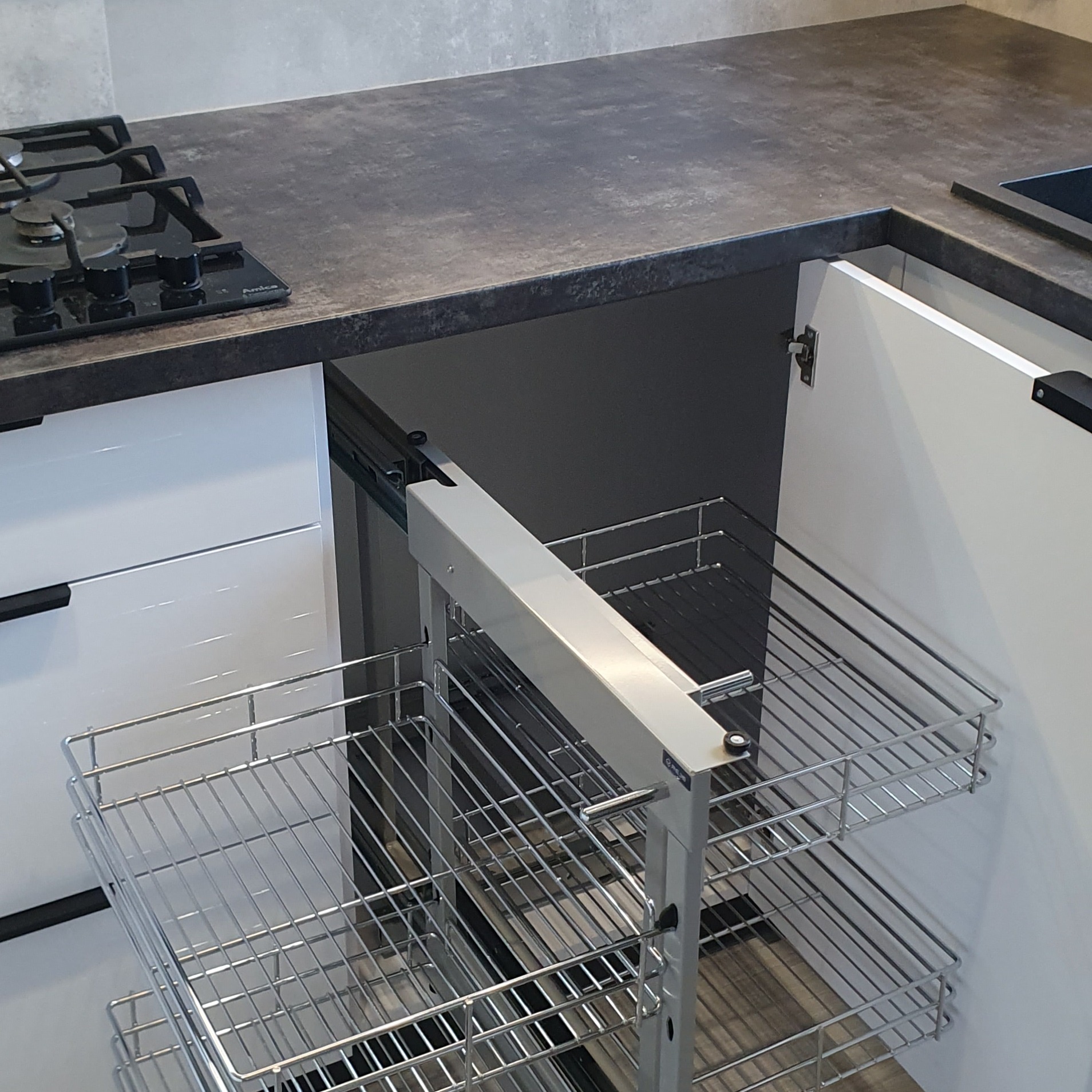 Wysuwany, metalowy system koszy cargo w szafce kuchennej z białymi frontami i blatem w ciemnym odcieniu szarości.