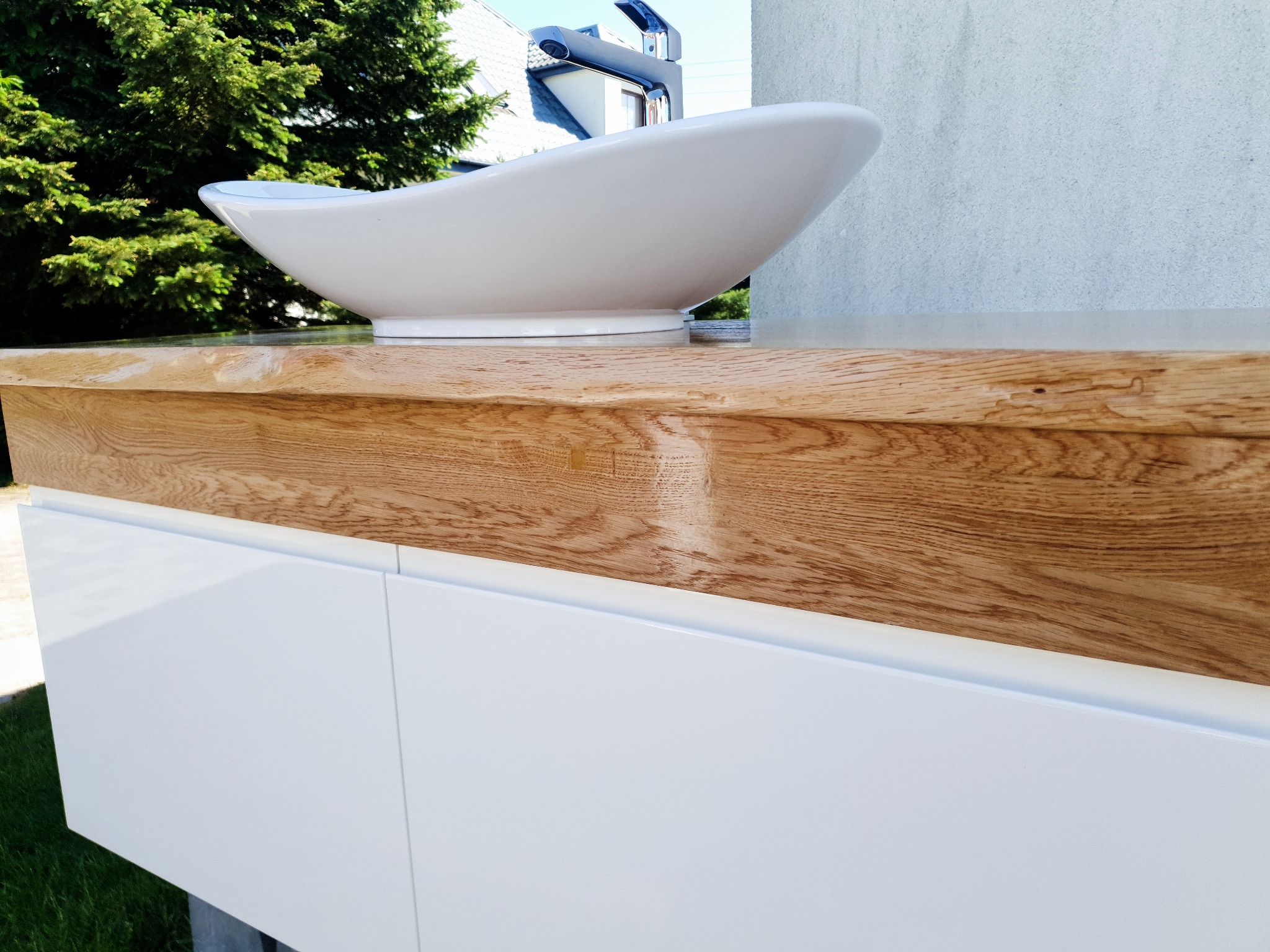Biała umywalka o owalnym kształcie zamontowana na blacie z jasnego drewna, pod którym znajdują się białe szafki z gładkimi frontami. W tle widoczne drzewa i fragment budynku.
