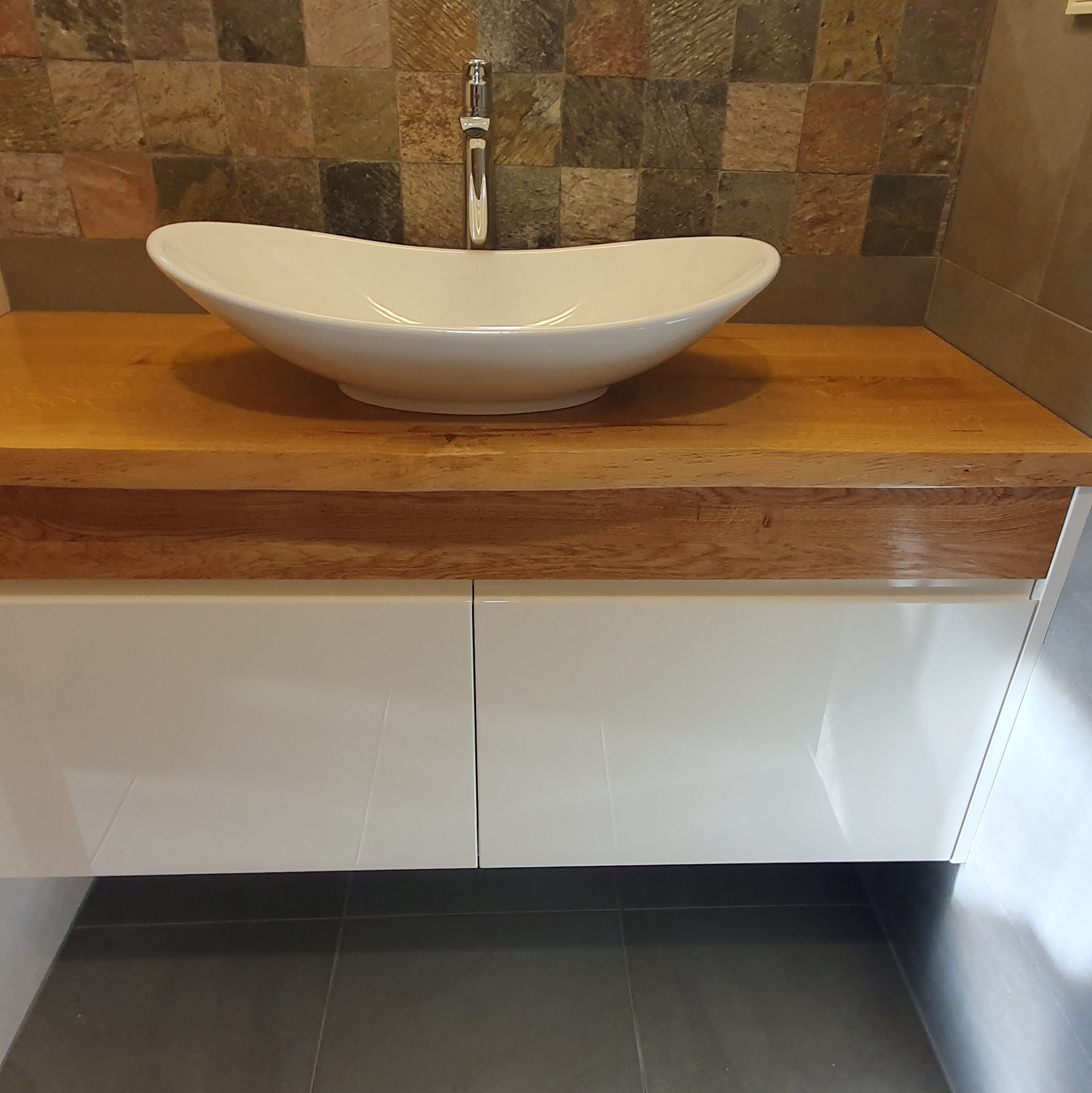 Biała, owalna umywalka nablatowa z chromowaną baterią na blacie z jasnego drewna, podwieszana szafka z białymi frontami, ściana wyłożona mozaiką z kamienia naturalnego.