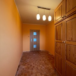 AROU Home Arkadiusz Nejman - Pomarańczowy pokój z drewnianą podłogą w jodełkę, starą szafą i drzwiami z trzema szybkami, oświetlony lampą z trzema kulistymi kloszami.