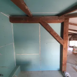 Montaż płyt gipsowo-kartonowych na poddaszu z widocznymi drewnianymi belkami konstrukcyjnymi i przygotowaną instalacją.