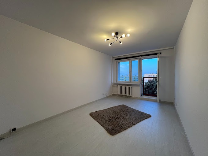 Pusty pokój po odświeżeniu, z białymi ścianami, szarym dywanem i nowoczesną lampą sufitową w stylu pająka, widok na balkon i okno z grzejnikiem.