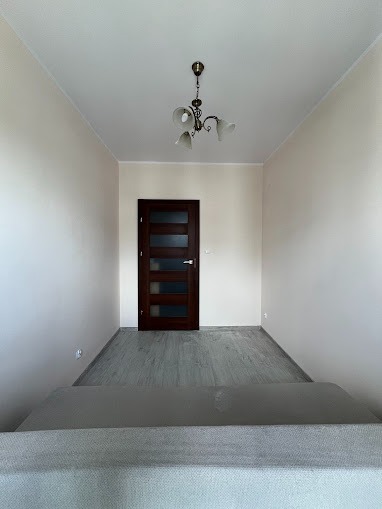 Pusty pokój po odświeżeniu: jasne ściany, szara podłoga imitująca drewno, ciemne drzwi z przeszkleniami i ozdobny, mosiężny żyrandol z białymi kloszami.