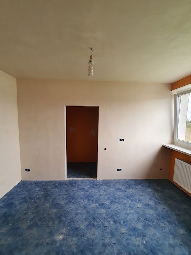 Pokój po szpachlowaniu, z widocznymi śladami po gładzi, gniazdkami elektrycznymi i niebieską podłogą imitującą morze, przez otwarte drzwi widać pomarańczową ścianę w drugim pomieszczeniu.