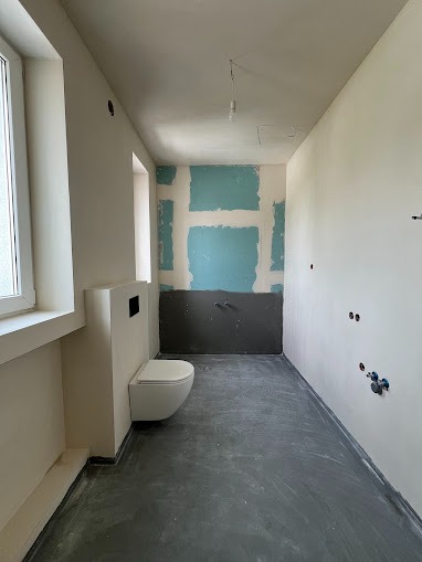 Wnętrze łazienki w trakcie prac wykończeniowych: zamontowana podtynkowa toaleta wisząca, fragment ściany z niebieskimi fragmentami płyt, przygotowywane instalacje, surowa podłoga pokryta wylewką.