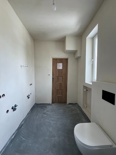 Wnętrze łazienki w trakcie prac wykończeniowych: ściany po szpachlowaniu, przygotowane do malowania, widoczne przyłącza wodno-kanalizacyjne, zamontowana toaleta podwieszana.