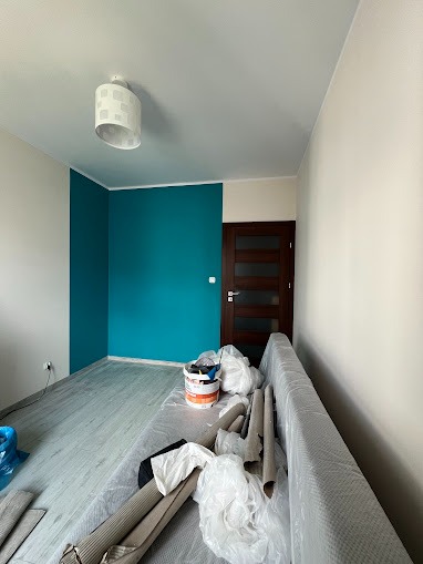 Świeżo pomalowany pokój z turkusową ścianą, zabezpieczona folią kanapa, puszki z farbą i wałki malarskie na podłodze.