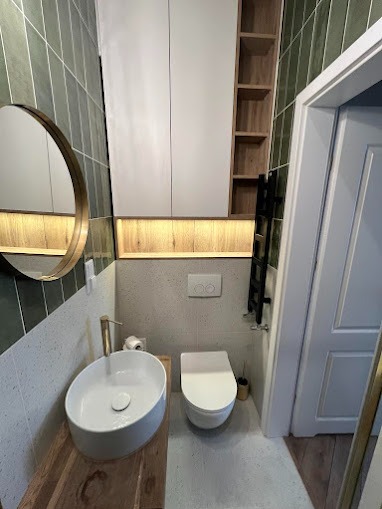 Nowoczesna łazienka z owalną umywalką nablatową na drewnianym blacie, podświetlane szafki wiszące, toaleta podtynkowa i zielone płytki na ścianach.