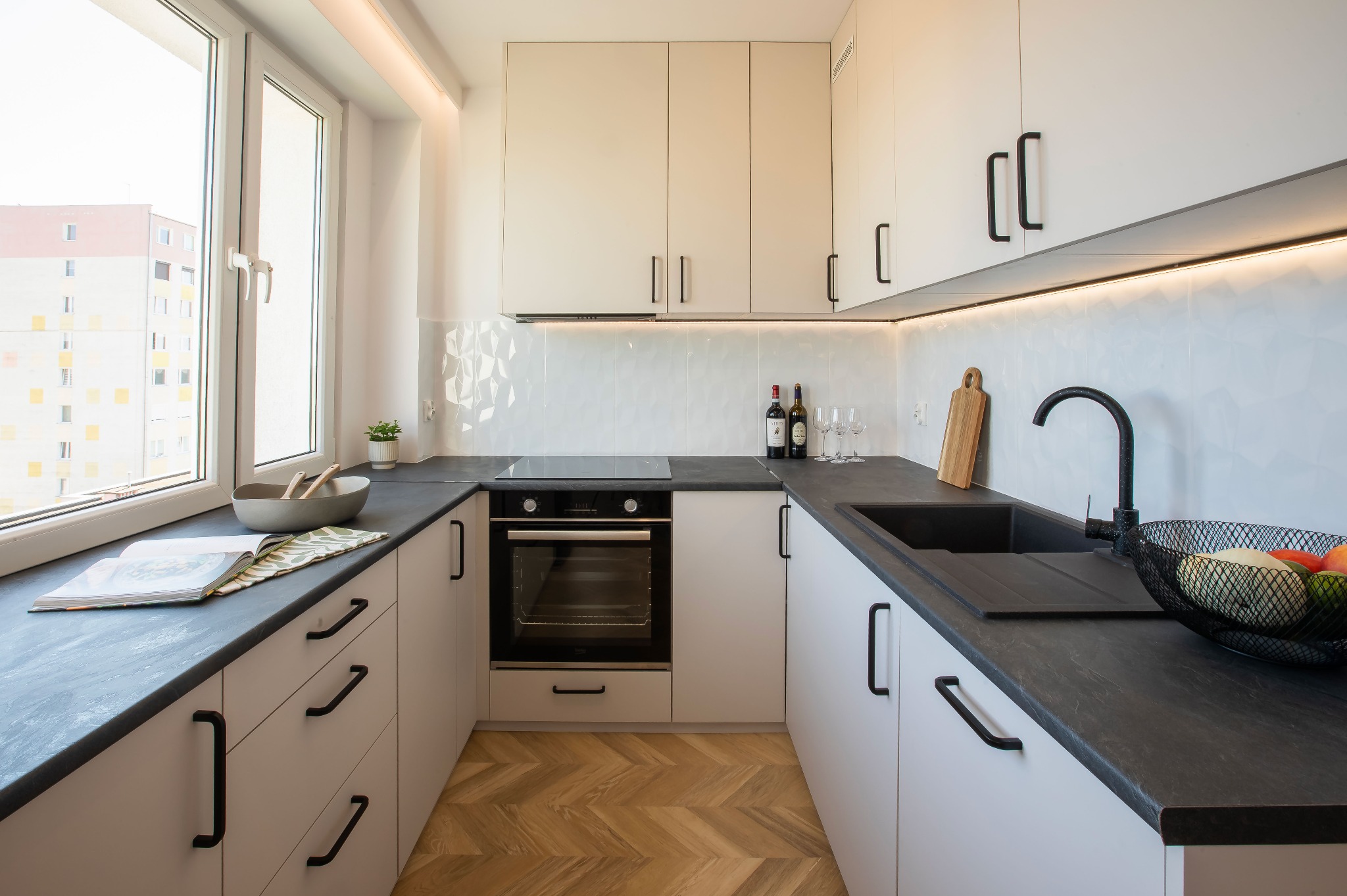 Nowoczesna kuchnia w bieli i czerni z drewnianą podłogą w jodełkę. Na blacie książka kucharska, owoce i wino. Minimalistyczny design z oświetleniem LED.