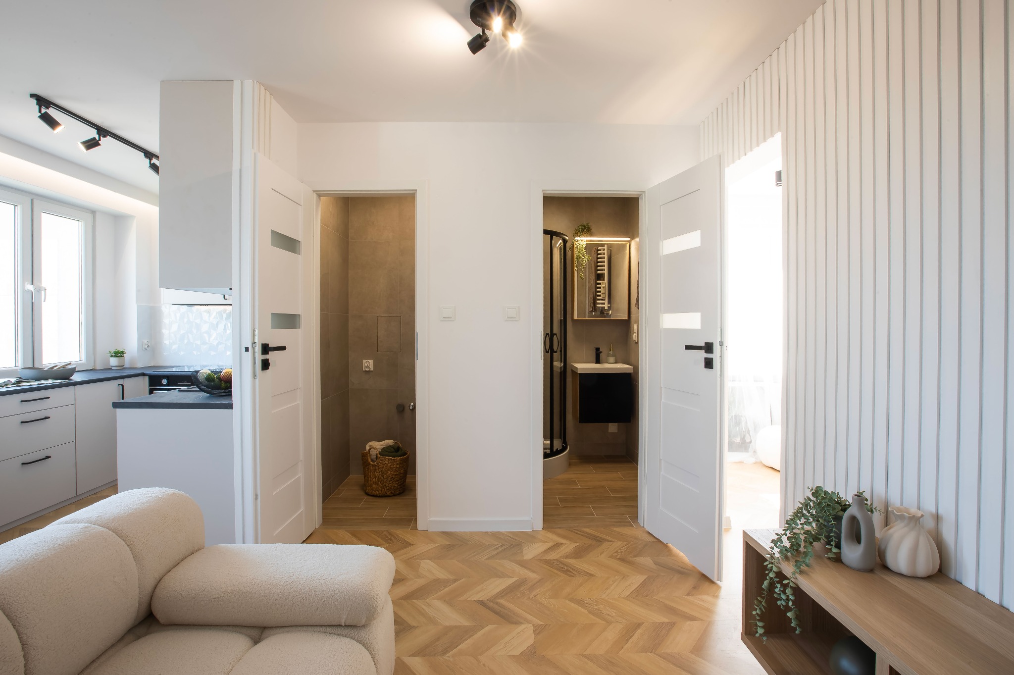Przestronne wnętrze po remoncie: widok na kuchnię, pralnię i łazienkę z parkietem w jodełkę. Białe ściany, drzwi z przeszkleniami i nowoczesne oświetlenie.