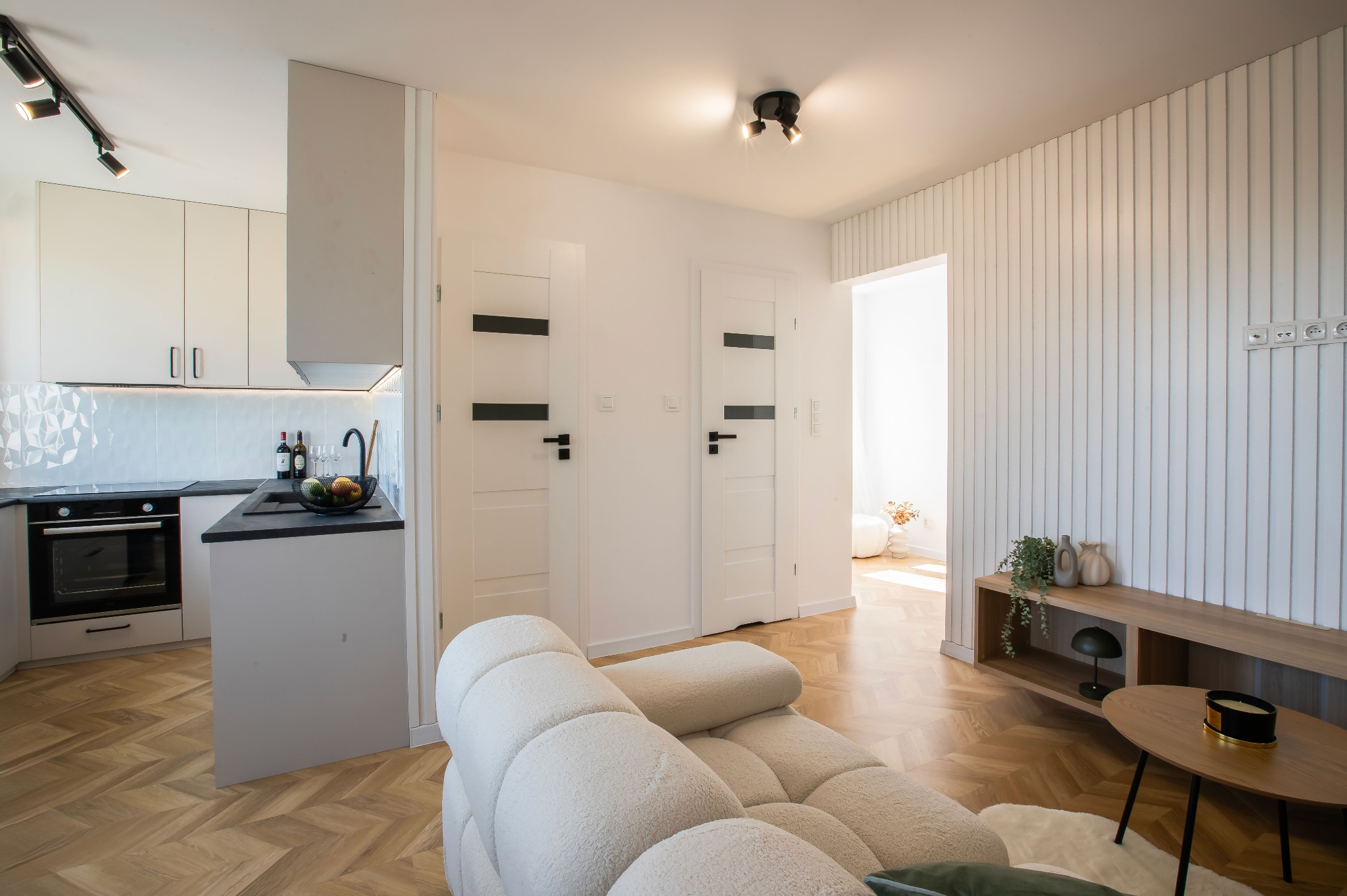 Jasne wnętrze po remoncie: parkiet w jodełkę, białe ściany z dekoracyjnymi panelami, nowoczesna kuchnia z czarnym blatem i minimalistyczne oświetlenie.