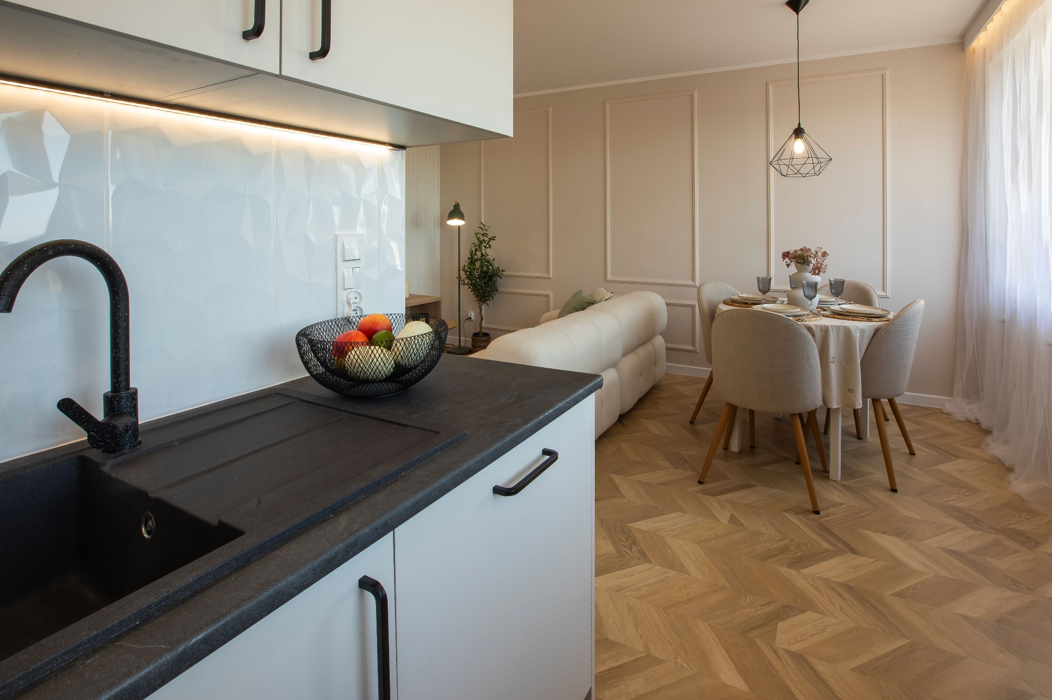 Nowoczesna kuchnia z czarnym zlewem i owocami w koszyku. W tle salon z jadalnią, parkiet w jodełkę i dekoracyjne panele ścienne. Jasne, przestronne wnętrze.