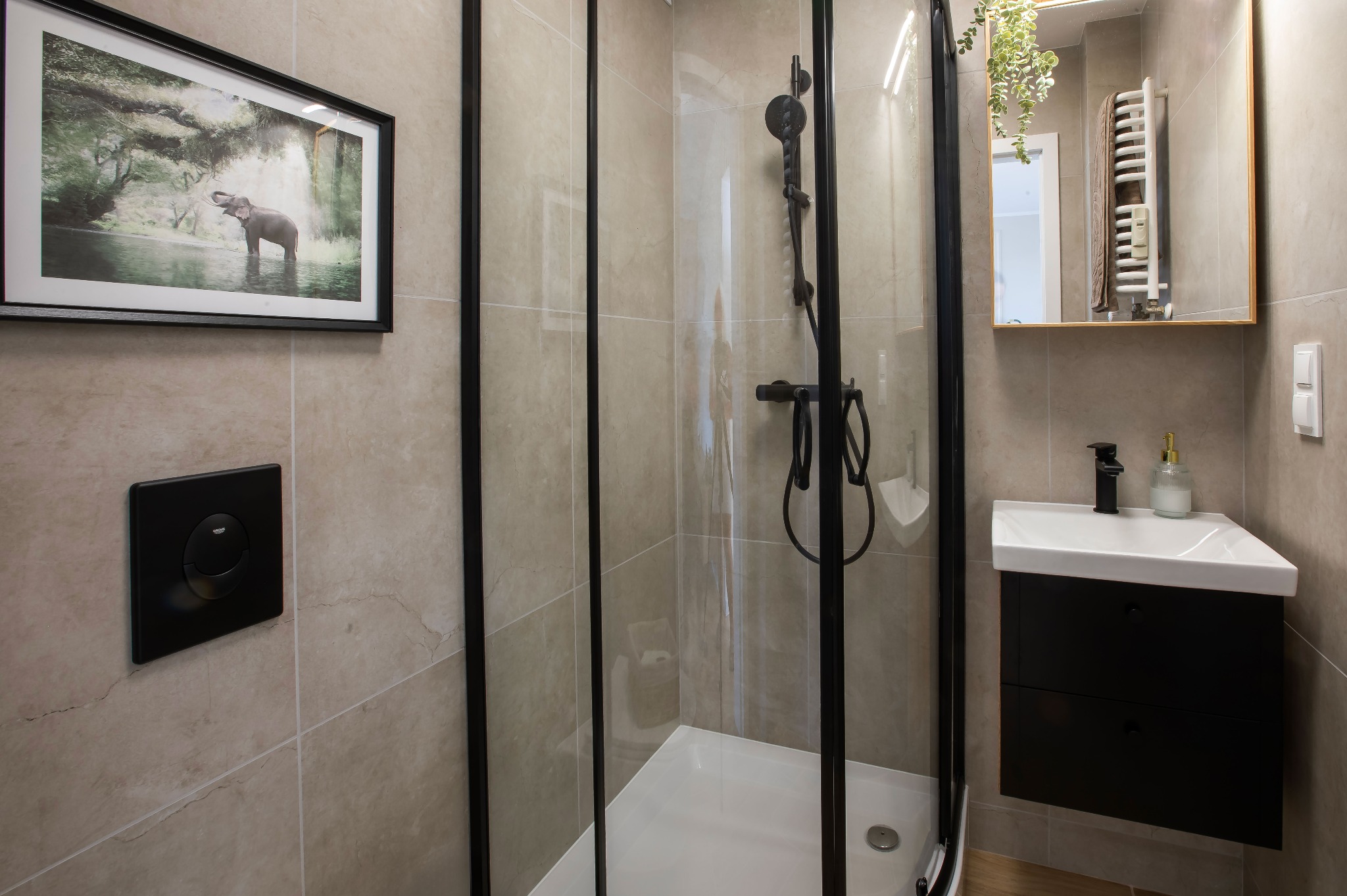Nowoczesna łazienka z kabiną prysznicową w czarnej ramie, umywalką na szafce i beżowymi płytkami. Na ścianie wisi obraz z motywem słonia.