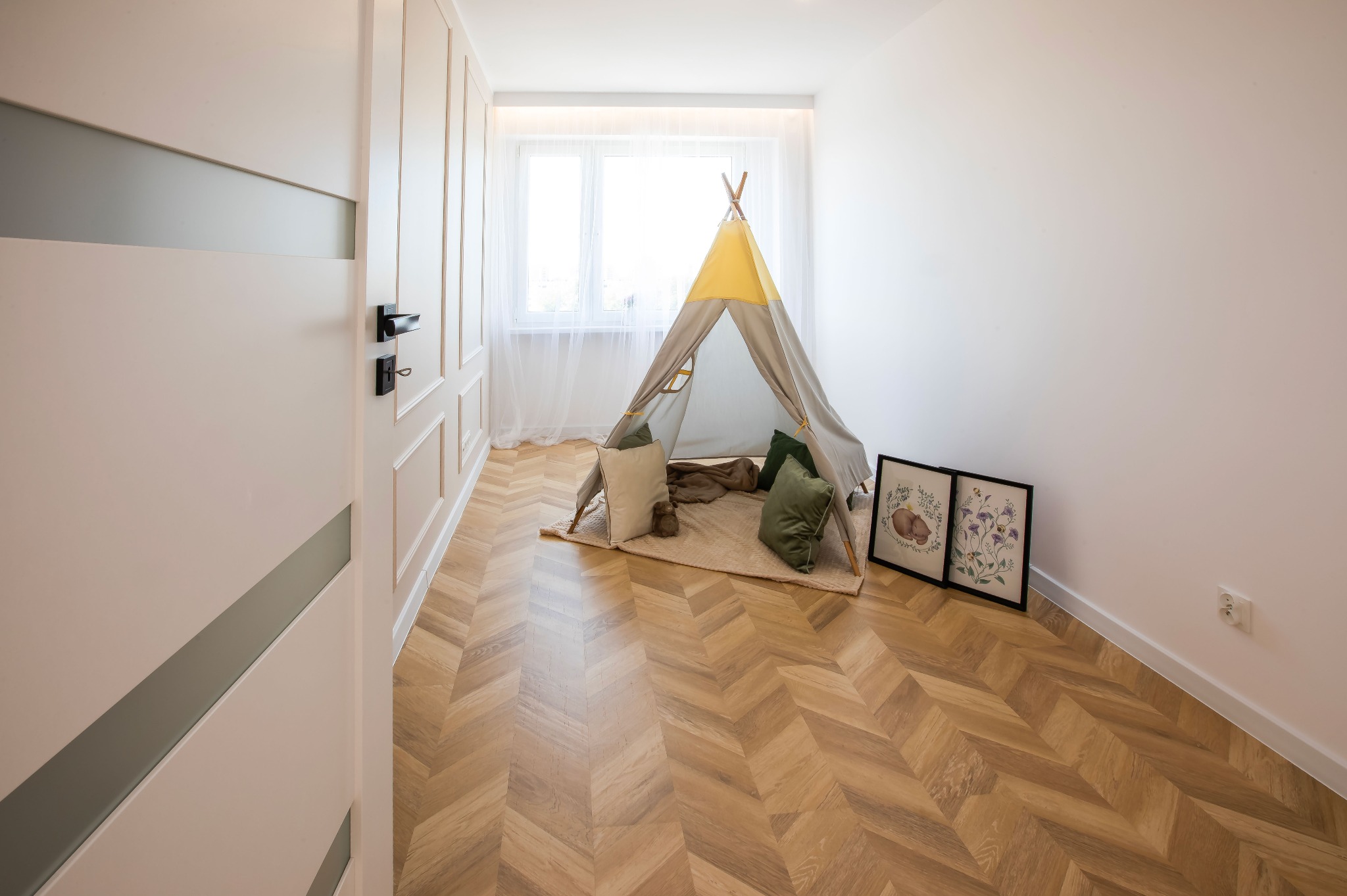Jasne wnętrze pokoju dziecięcego z parkietem w jodełkę, tipi i dekoracjami ściennymi. Widoczne drzwi z nowoczesnym designem i subtelne oświetlenie.