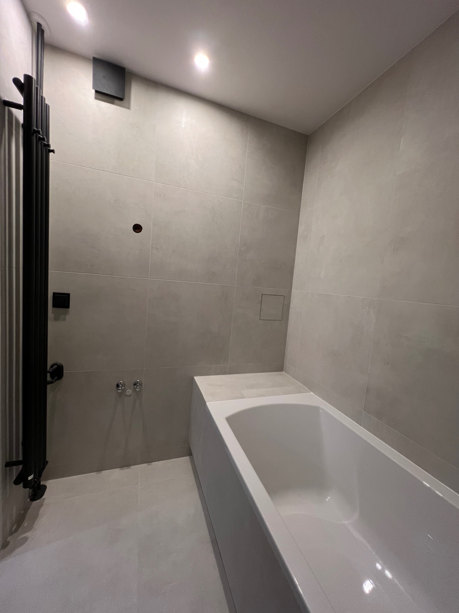 Wykończona łazienka z szarymi płytkami, białą wanną i czarnym grzejnikiem drabinkowym. Minimalistyczny design, widoczne punkty świetlne na suficie.