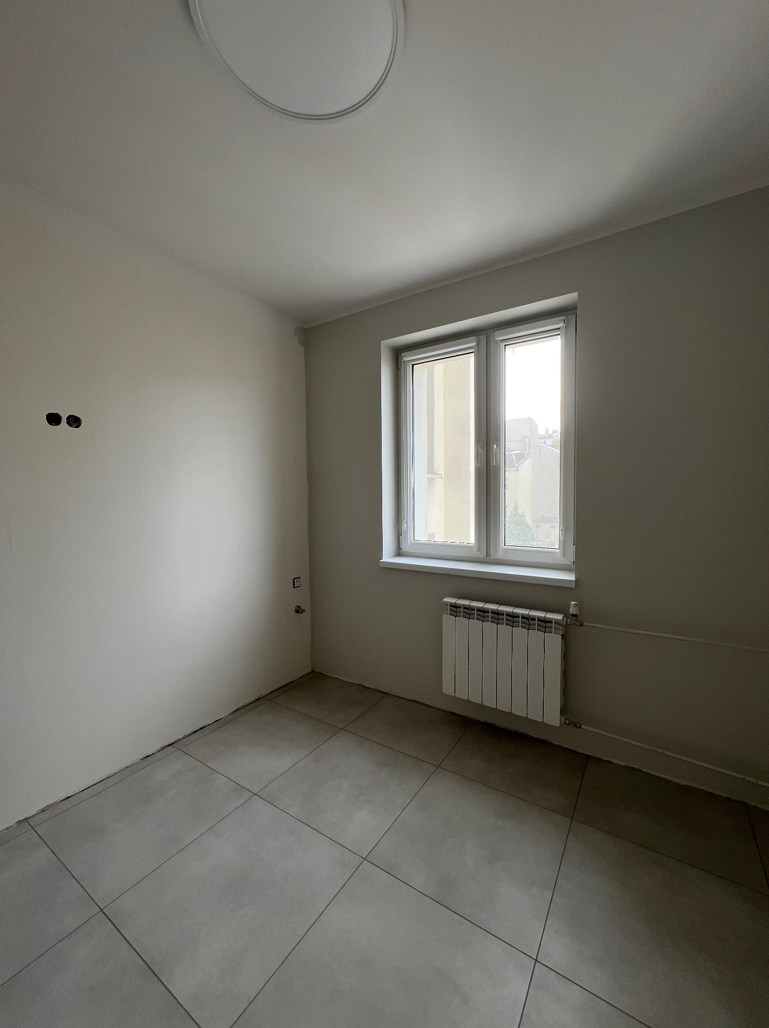 Wnętrze pokoju po remoncie: szare płytki podłogowe 60x60, świeżo pomalowane ściany, nowy grzejnik pod oknem. Widoczne gniazdka elektryczne i instalacja.