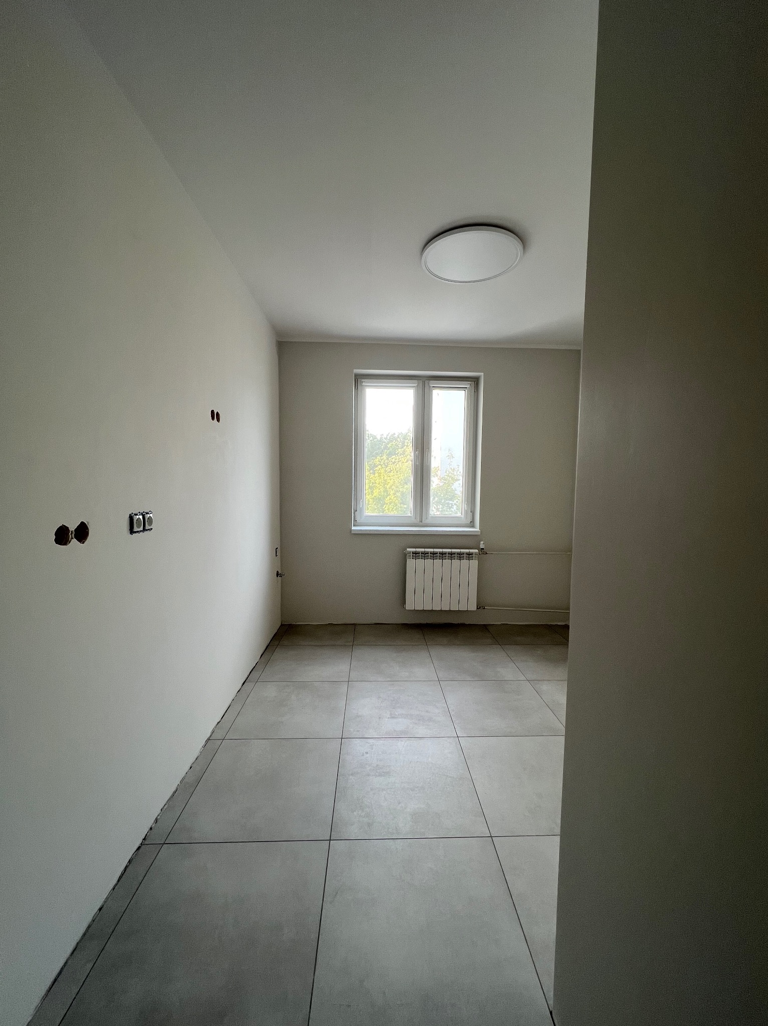 Jasne wnętrze po remoncie: świeżo pomalowane ściany, ułożona podłoga z płytek, okno z grzejnikiem. Widoczne puszki elektryczne. Minimalistyczny design.
