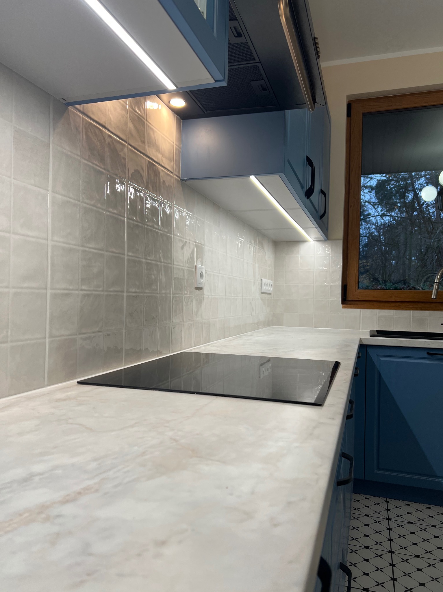 Wykończona kuchnia z płytkami na ścianie, marmurowym blatem i niebieskimi szafkami. Widoczne oświetlenie LED pod szafkami i okno z drewnianą ramą.