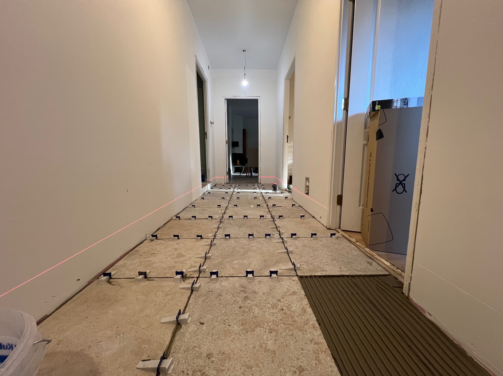 Korytarz w trakcie układania płytek, widoczne poziomice laserowe i system poziomowania płytek. Częściowo ułożone płytki z klejem. Remont w toku.