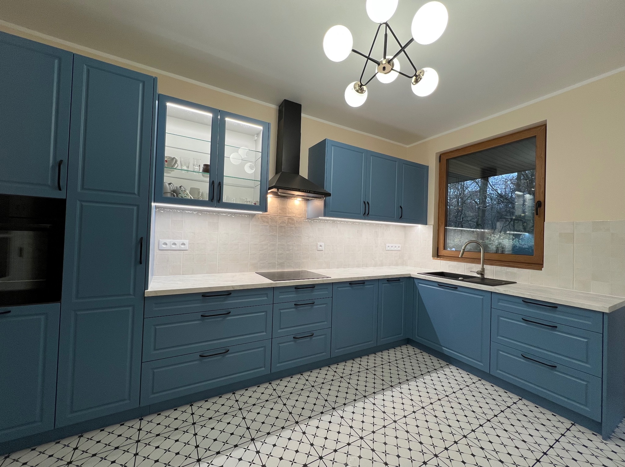 Niebieska, narożna zabudowa kuchenna z oświetleniem LED podszafkowym i wzorzystą podłogą. Nowoczesny design z czarnym okapem i lampą sufitową.